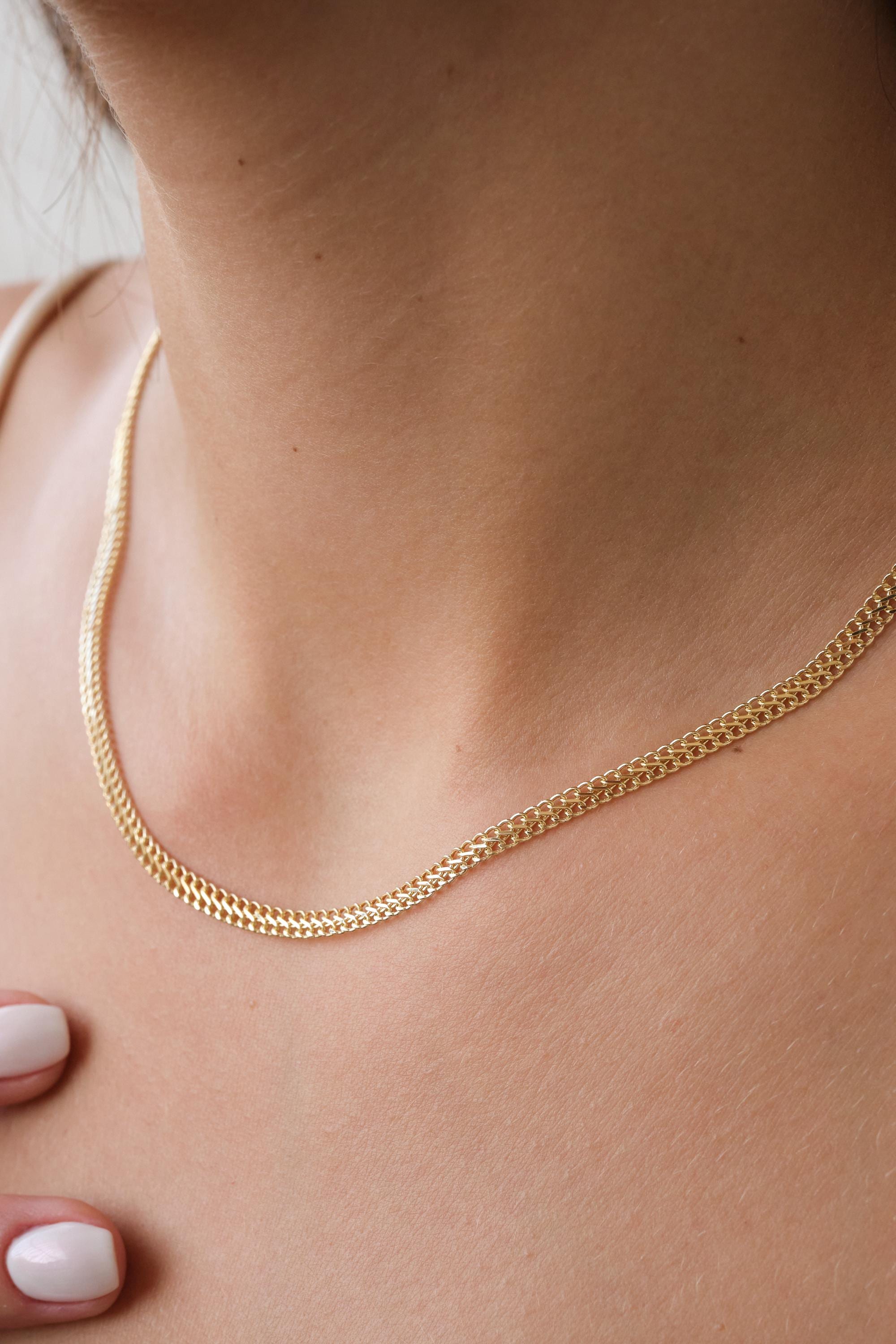May include: A gold-colored chain necklace with a delicate, woven design. The necklace is displayed on a person's neck, showcasing its simple yet elegant style. The chain's links create a textured surface, reflecting light.