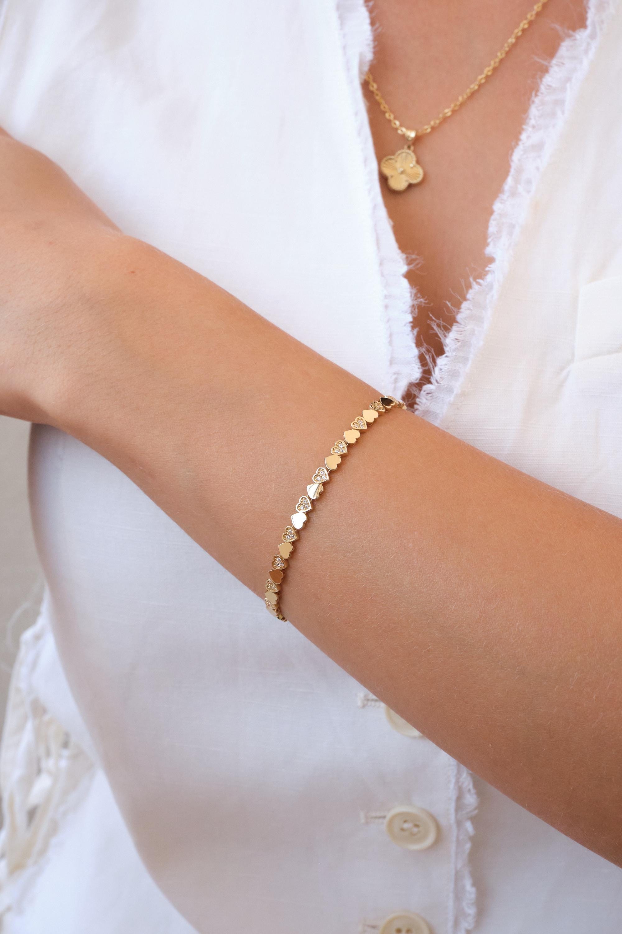 May include: A gold bracelet with heart-shaped links and small diamond accents. The bracelet is worn on a person's wrist. A gold necklace with a clover pendant is also visible.