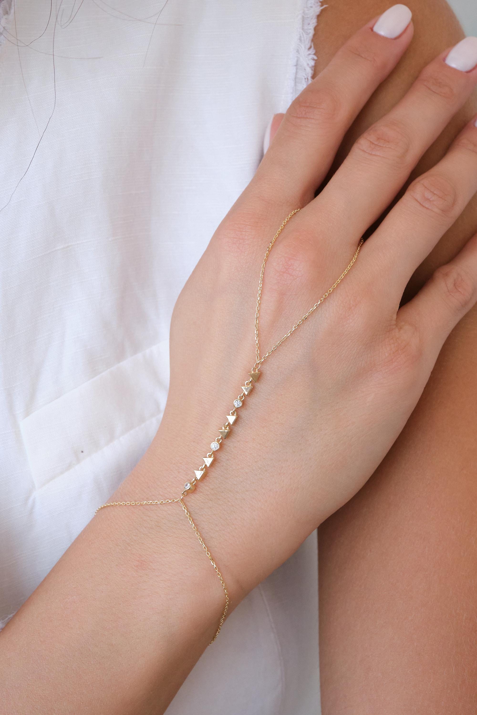 May include: A delicate gold hand chain bracelet. The bracelet features a series of small triangle and round-shaped charms, some with clear gemstones. The chain connects to a ring and wraps around the wrist and hand.