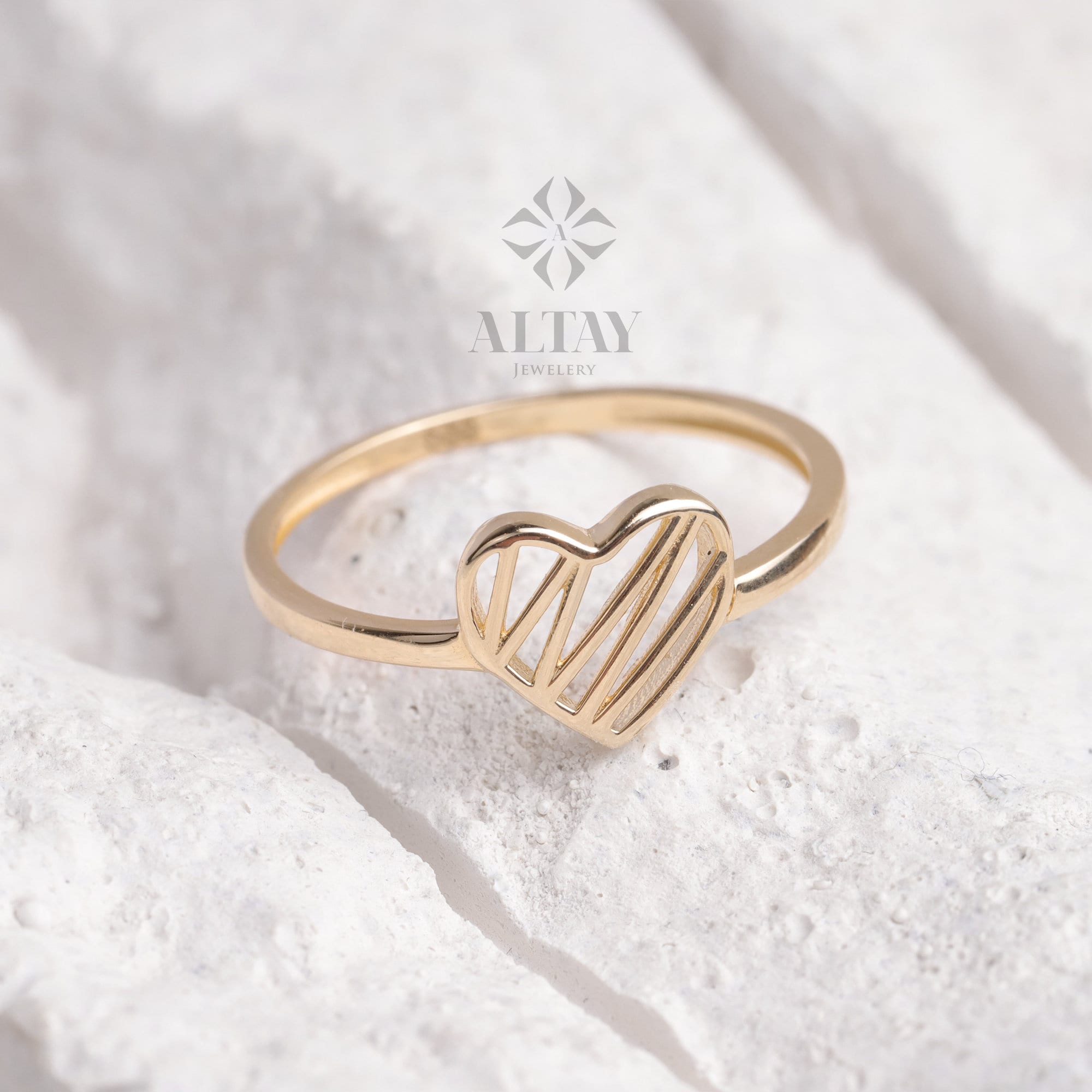 May include: A gold ring with a heart-shaped design. The heart features a series of parallel lines, creating an open, airy look. The ring is set against a white, textured background, with the "ALTAY JEWELLERY" logo visible.