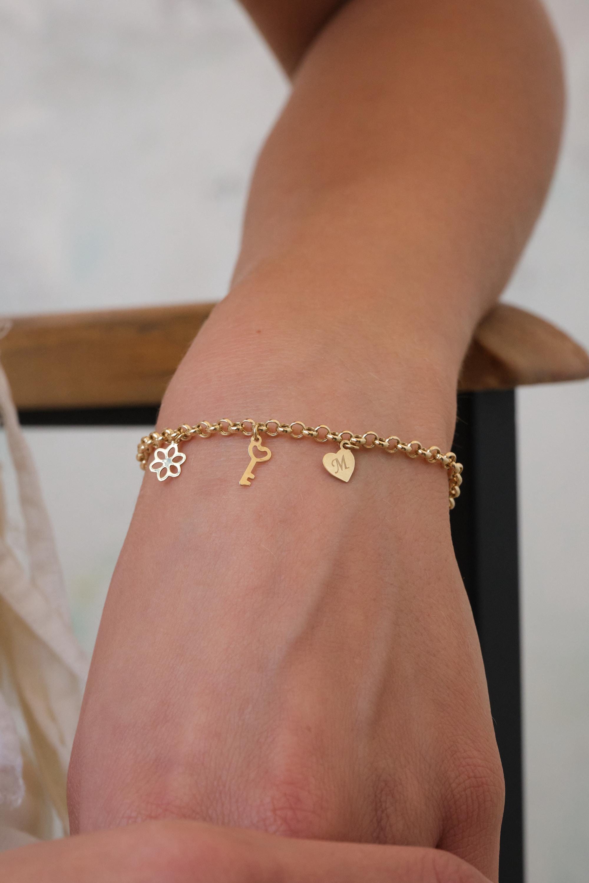 May include: Gold-toned charm bracelet with a flower, key, and heart charm. The heart charm has the letter "M" engraved on it. The bracelet is worn on a wrist.