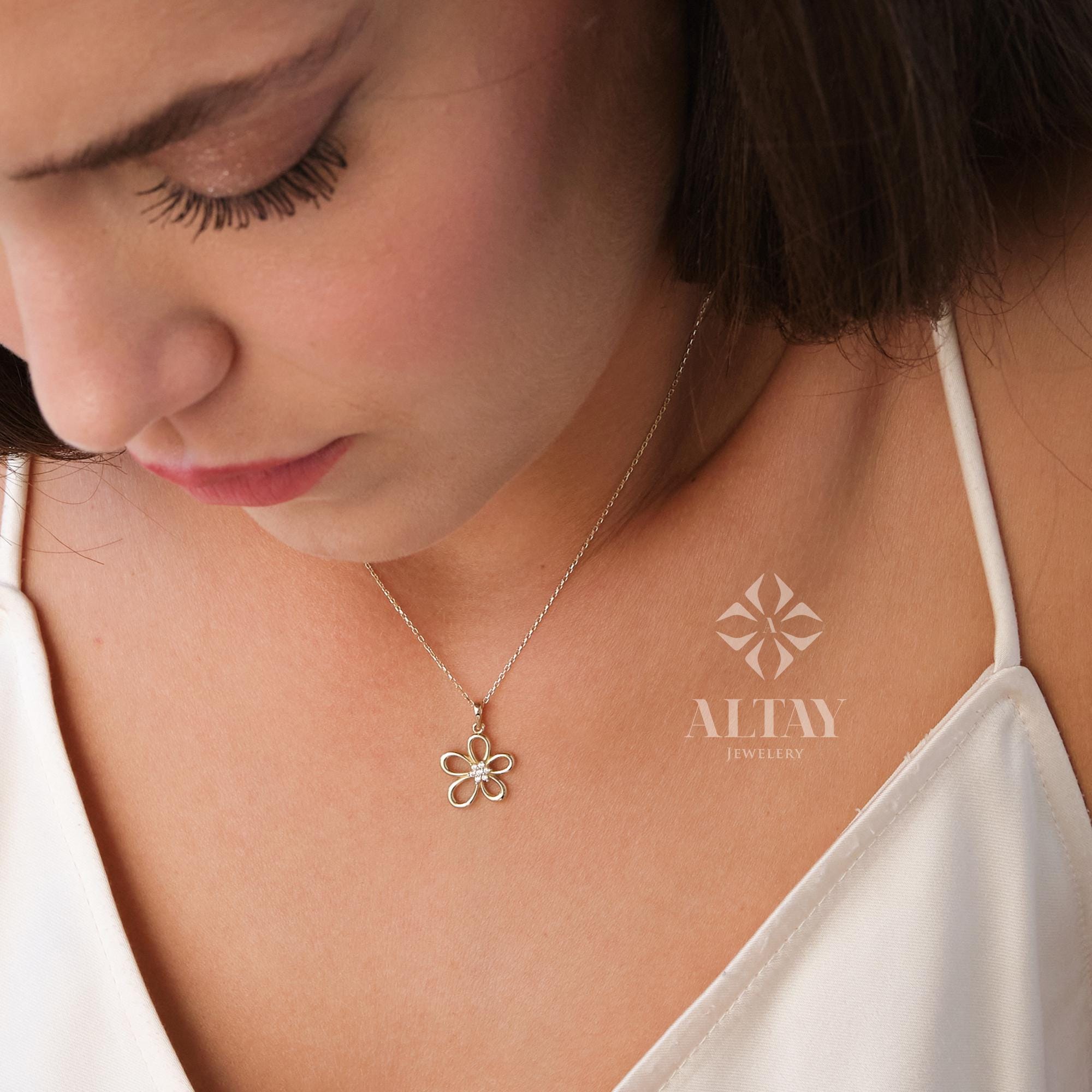 May include: A delicate gold necklace featuring a flower pendant with a small gemstone center. The necklace is worn on a person's neck, and the pendant is centered. The necklace is from Altay Jewelry.
