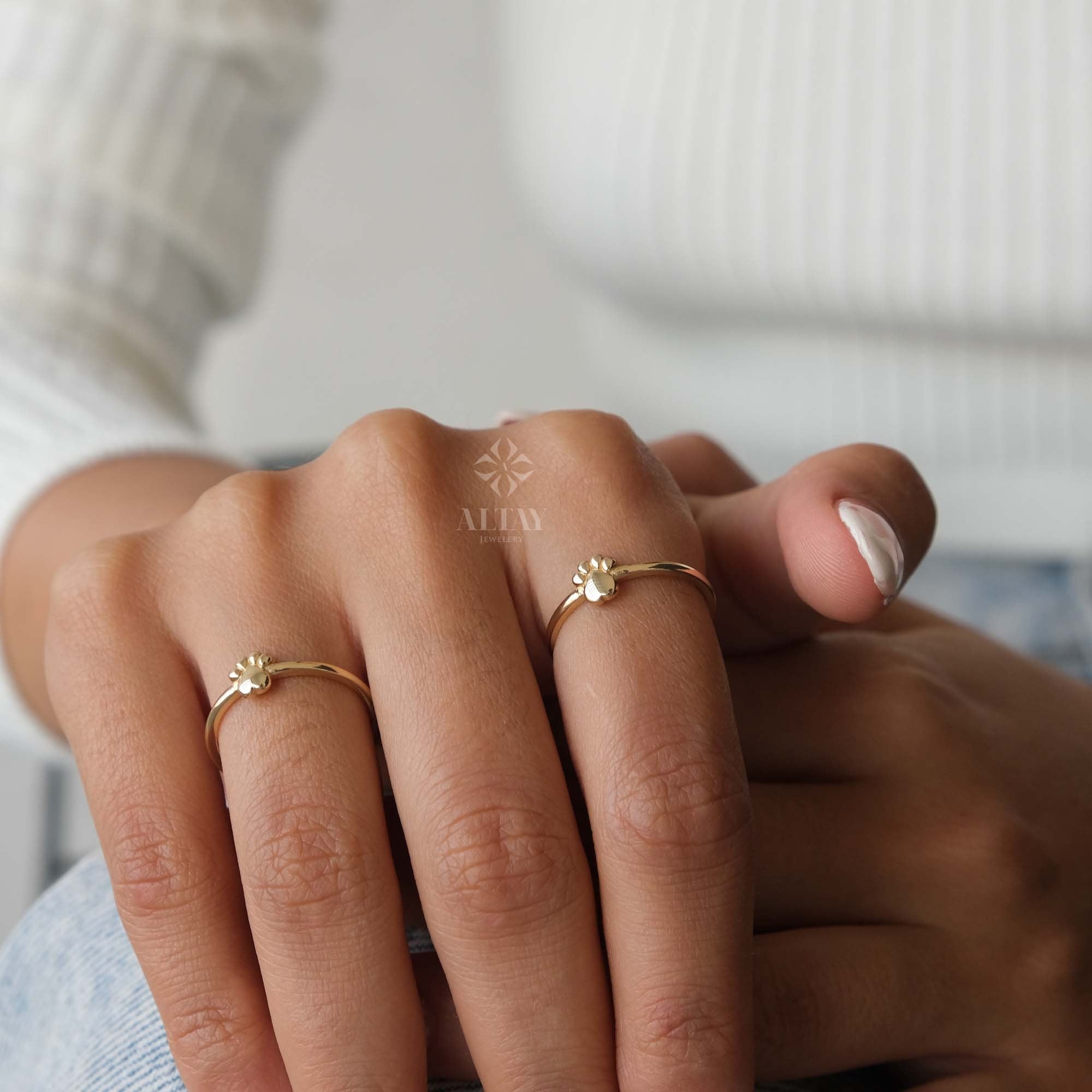 May include: Two gold rings with a paw print design. The rings are worn on the fingers of a person's hand.