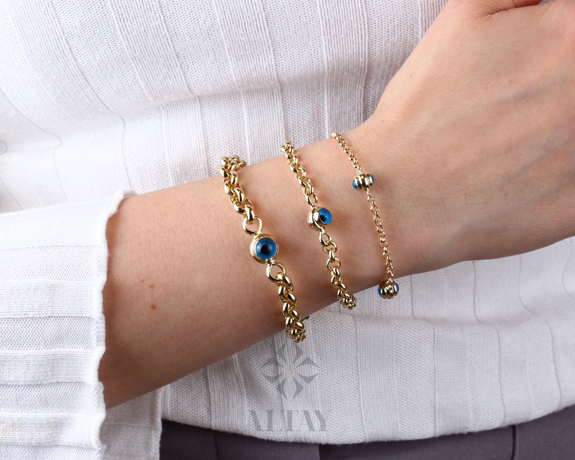 May include: Three gold chain bracelets with blue evil eye charms. The bracelets are layered on a person's wrist.