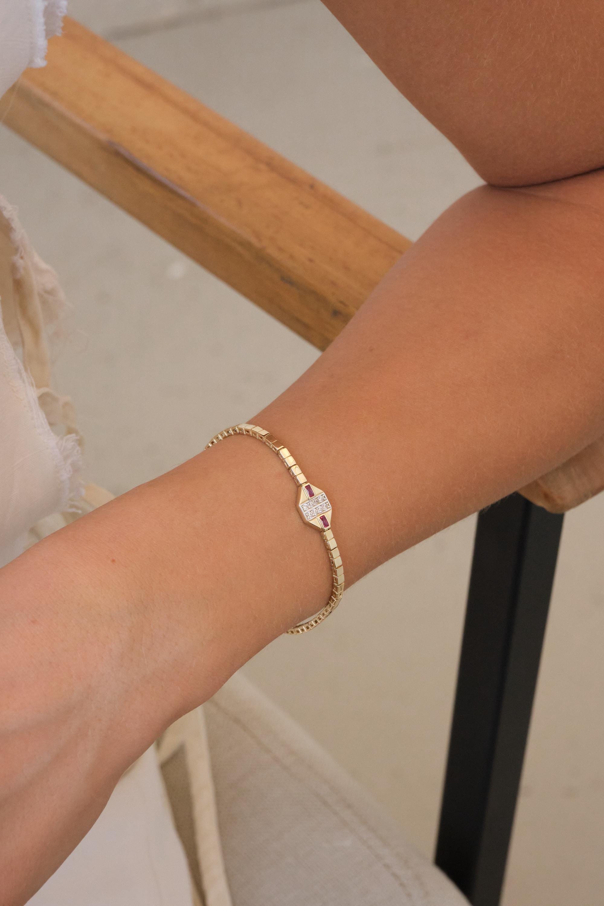 May include: A gold bracelet with a rectangular centerpiece. The centerpiece is adorned with small white and red gemstones. The bracelet is on a wrist.