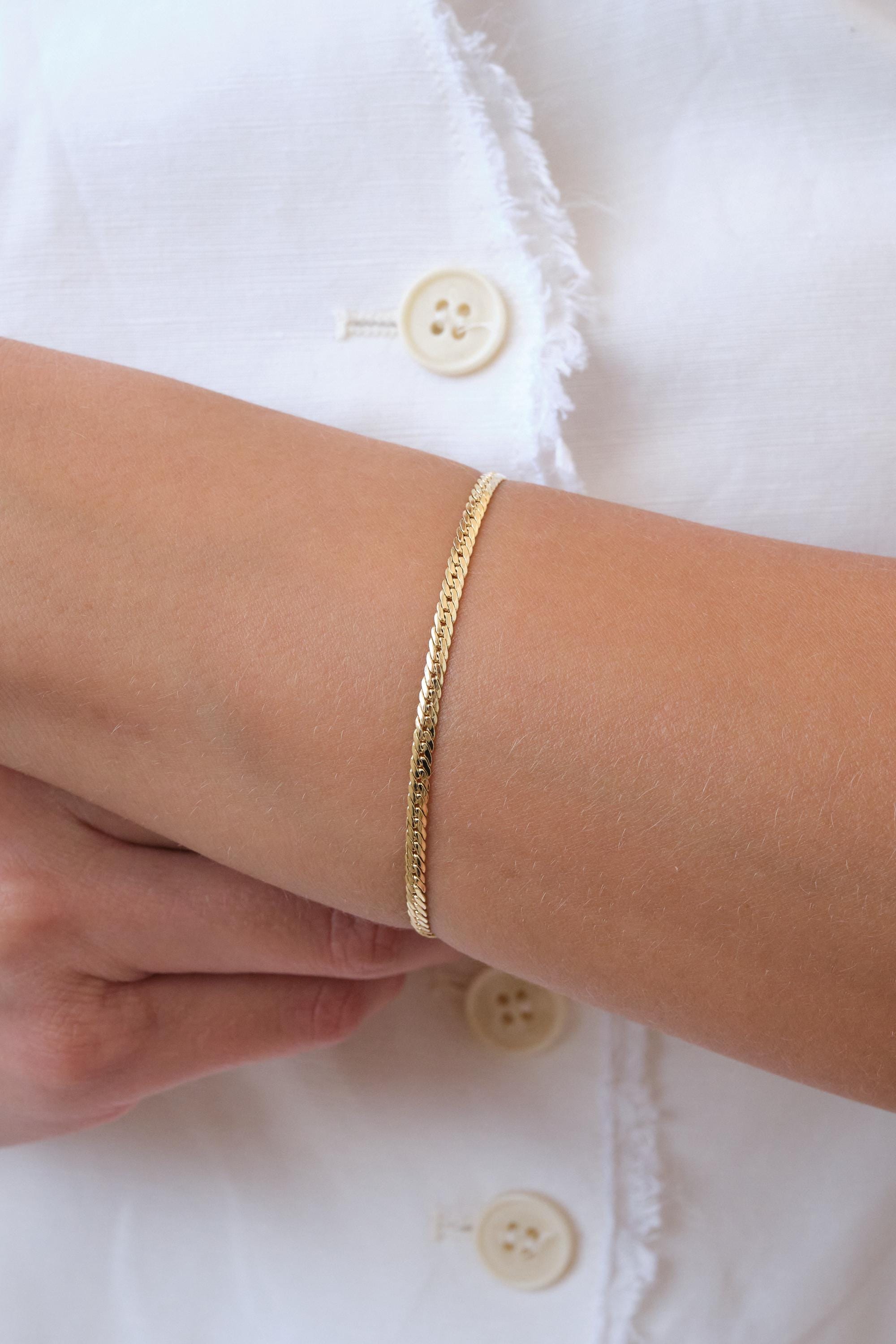 May include: A gold-colored herringbone bracelet. The bracelet is a thin, flat chain with a woven pattern. The bracelet is worn on a person's wrist. The background is a white shirt with a button.