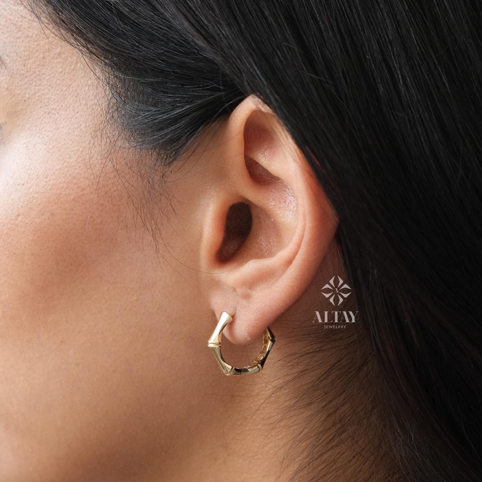 May include: Gold bamboo hoop earrings. The earrings are a simple design with a slightly rounded shape. The earrings are shown on a person's ear.