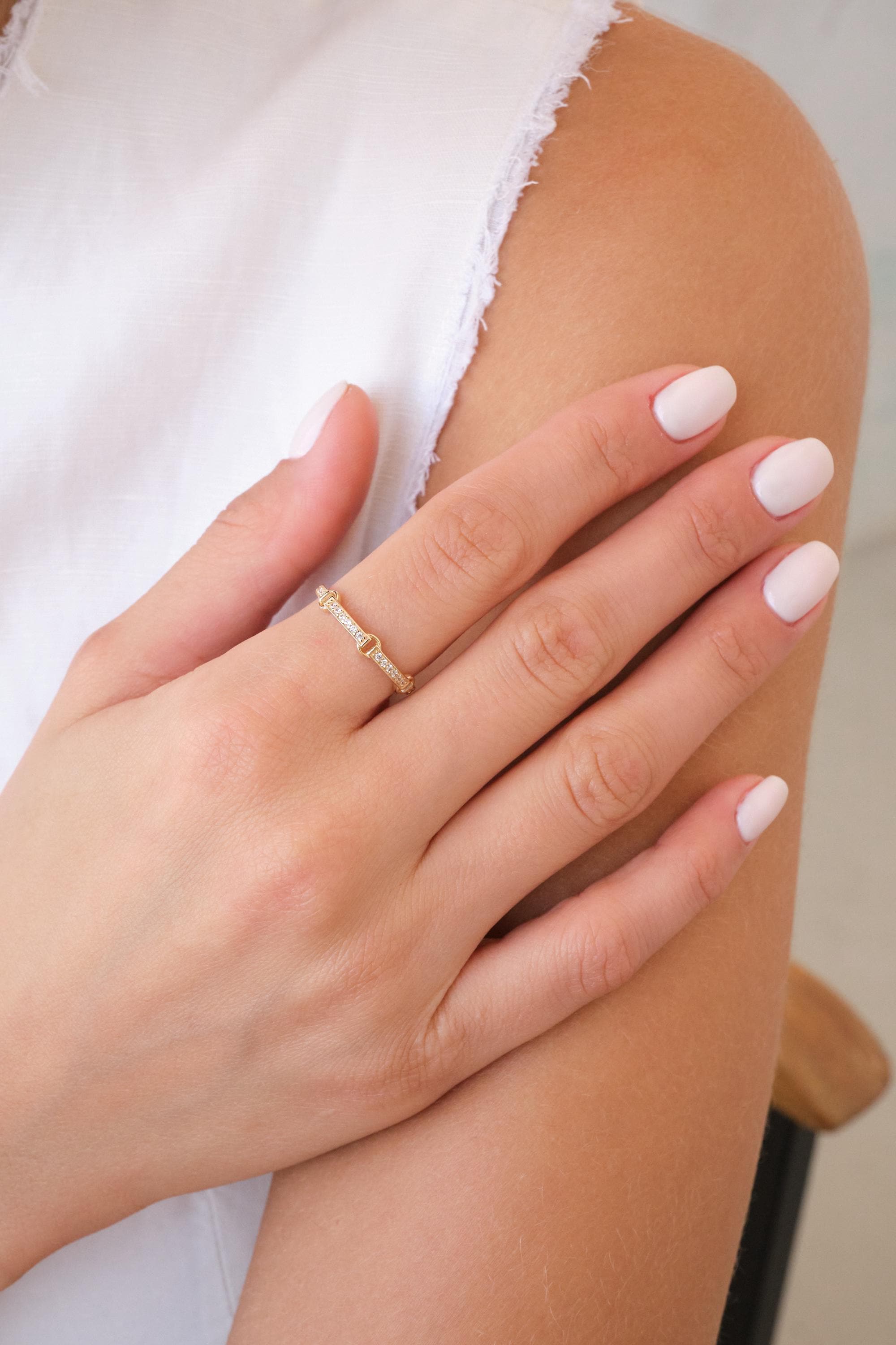 May include: A gold ring with a small heart-shaped design and embedded clear stones. The ring is on a person's finger, with a white top in the background. The ring is a delicate piece of jewelry.