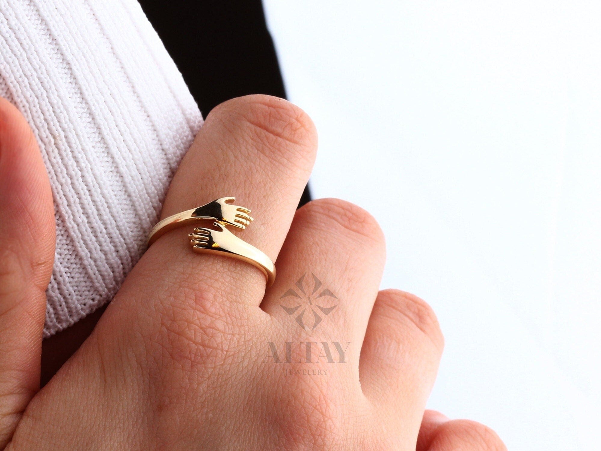 May include: Gold ring with two hands clasped together, symbolizing an embrace. The ring is worn on a finger.