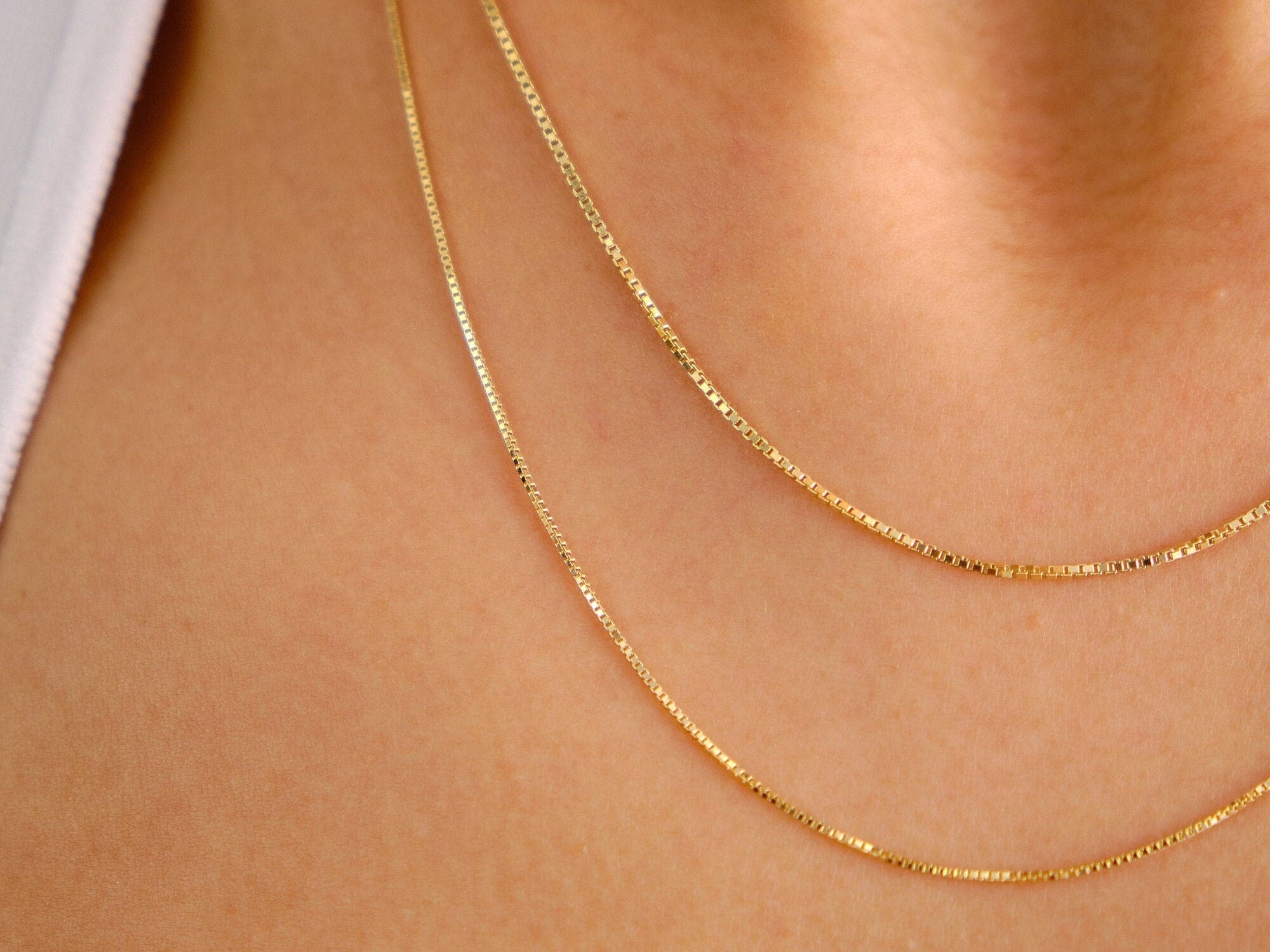 May include: Three gold chain necklaces layered on a person's neck. The chains are thin and delicate, with a box chain style.