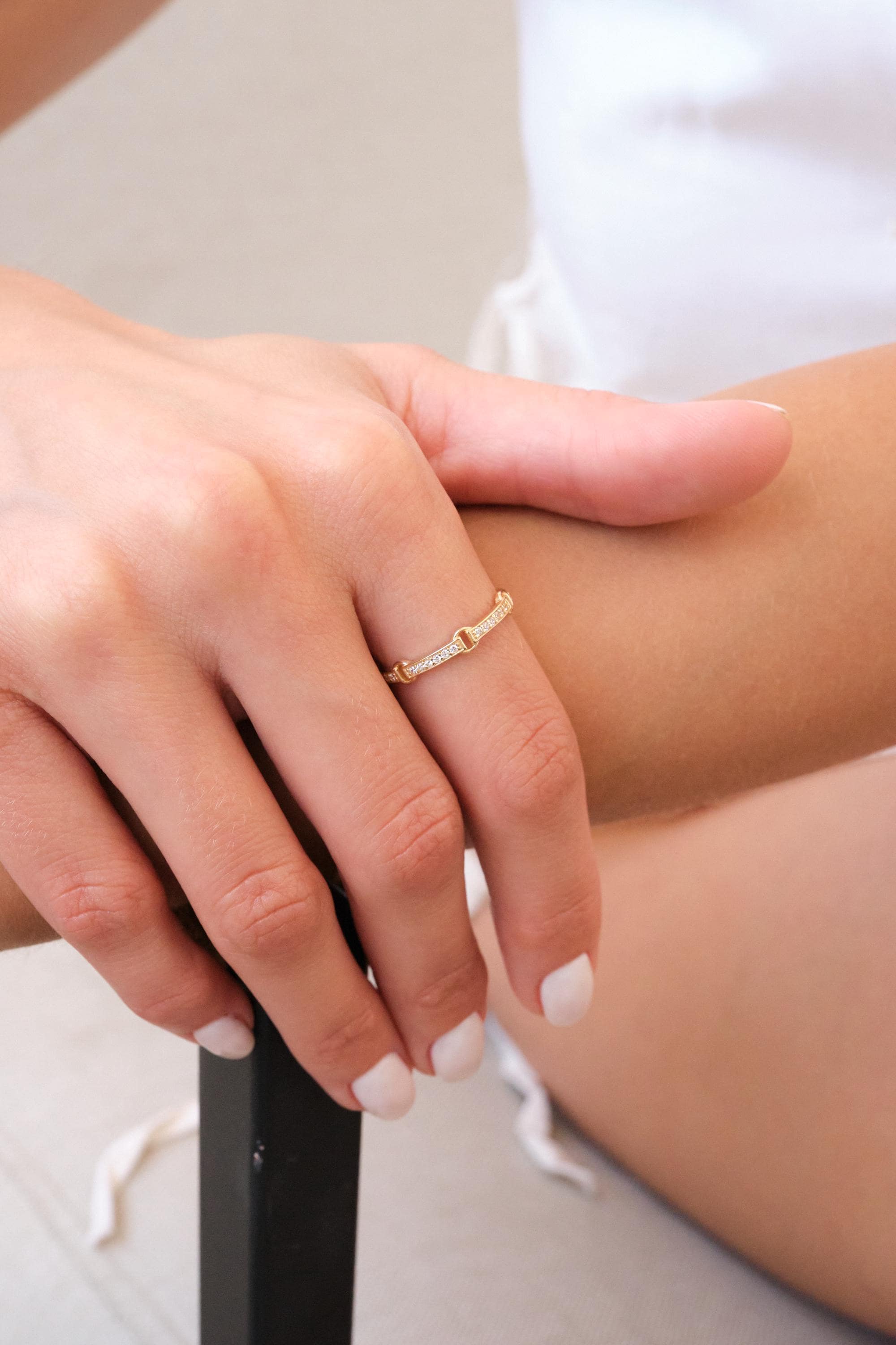 May include: A gold-colored ring with small, clear gemstones set along the top. The ring is on a person's finger, with the hand resting on a surface. The ring has a unique design, with a series of connected links.