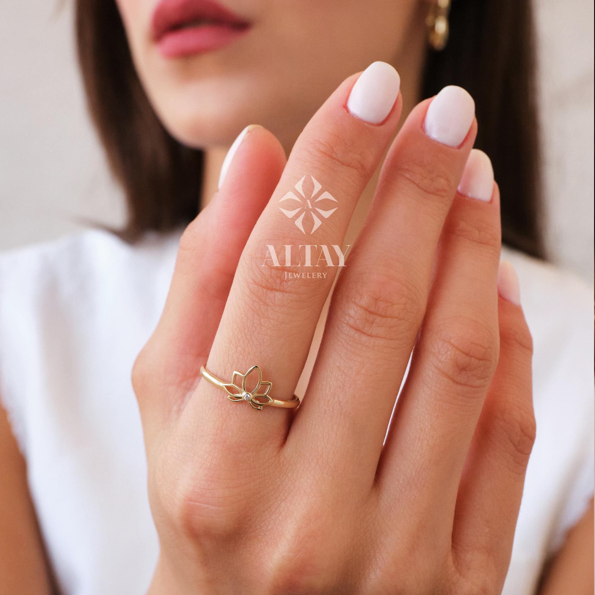 May include: A gold ring featuring a lotus flower design. The ring is delicate and has a thin band. The lotus flower is the focal point, with detailed petals. The ring is worn on a finger. The brand name "ALTAY JEWELLERY" is visible.