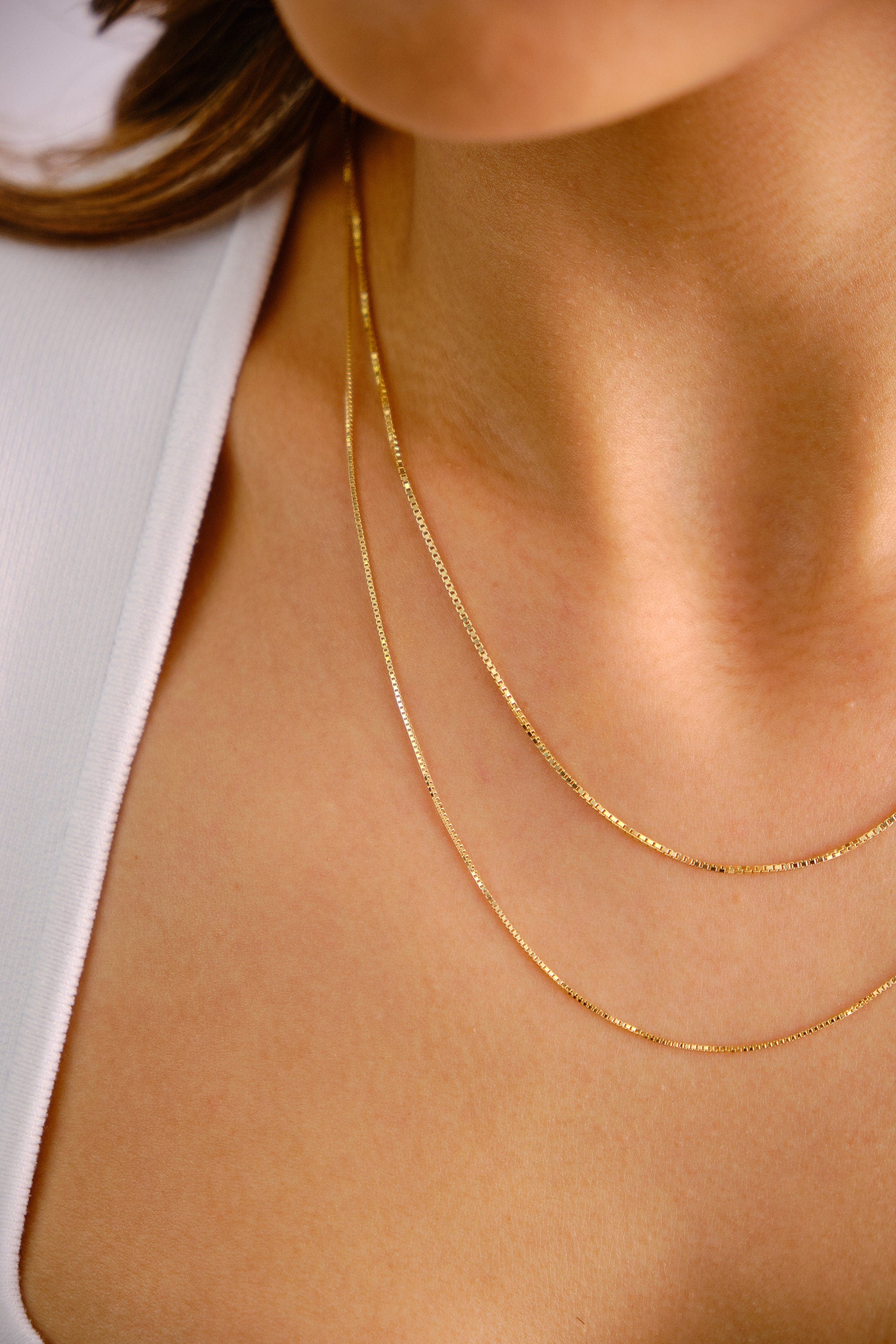 May include: Two gold chain necklaces layered on a person's neck. The chains are thin and delicate, with a box chain style.