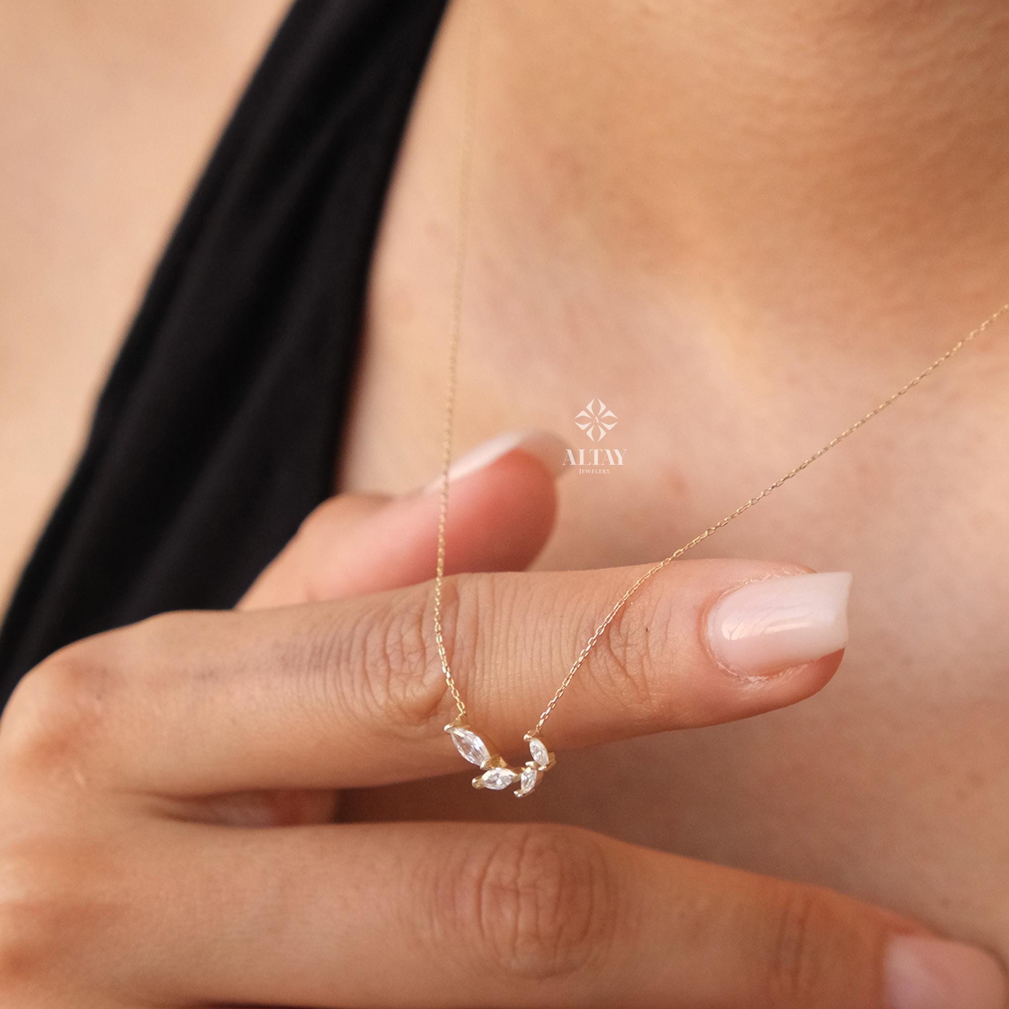 May include: A delicate gold necklace with a pendant featuring a row of small, clear marquise-shaped stones set in a curved design. The necklace is shown on a person's hand.