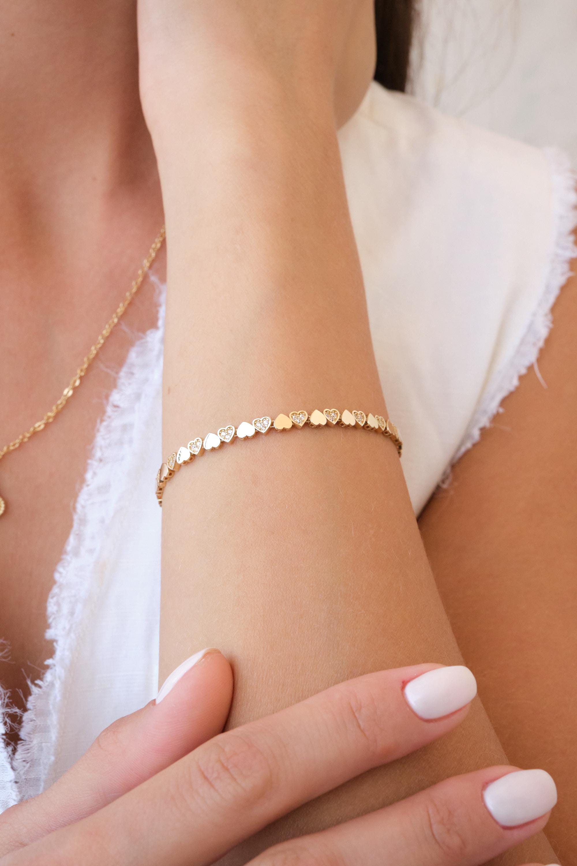 May include: A gold-colored bracelet featuring a series of heart-shaped charms. Some hearts have small, clear embellishments. The bracelet is worn on a wrist, and a gold necklace is also visible. The background is a white top.