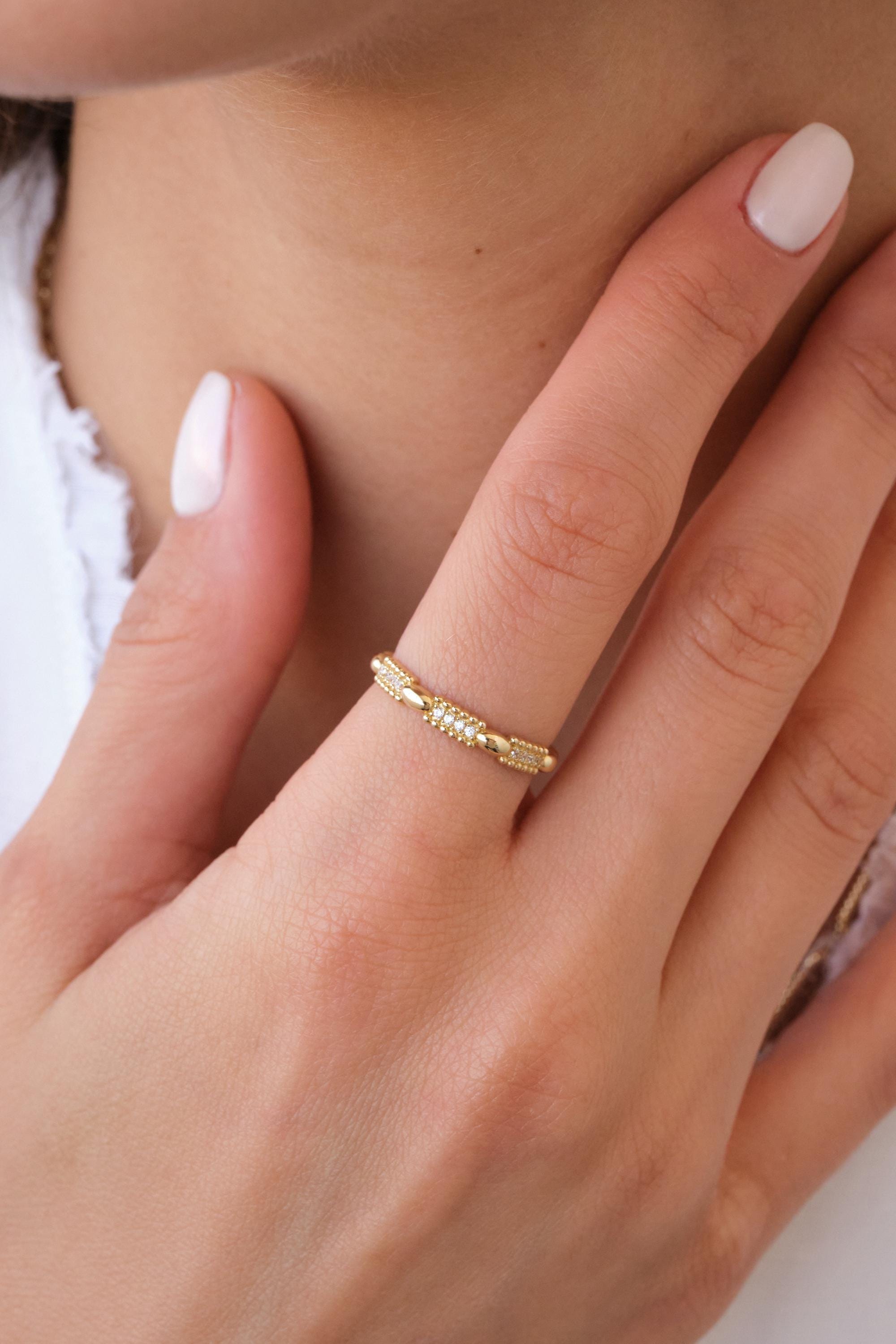 May include: A gold ring with a repeating pattern of small diamonds and textured gold segments. The ring is worn on a finger, with the hand resting near a person's neck. The ring's design is delicate and elegant.